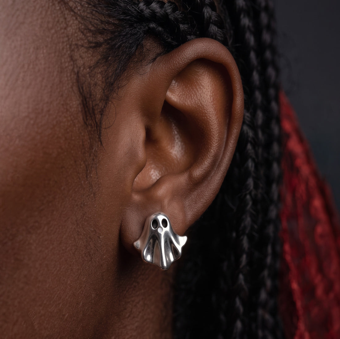 Silver Boo stud earrings worn on the ear, a silver ghost with black eyes against braids with red ends