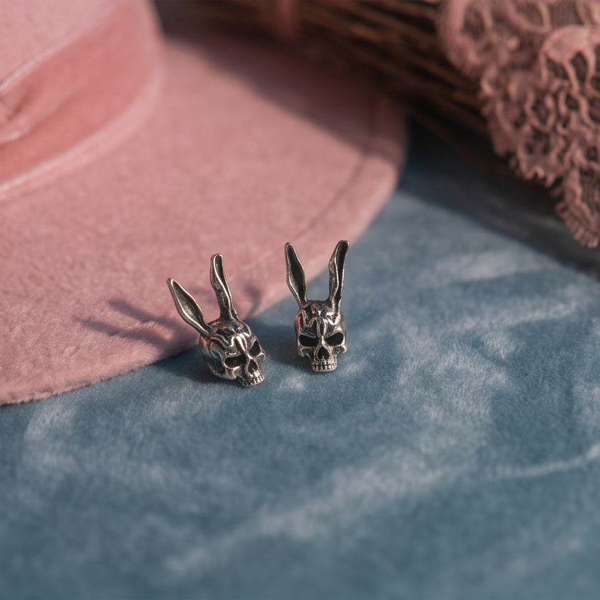 Darko Rabbit Skull stud earrings pair on blue velvet beside a pink felt hat and lace