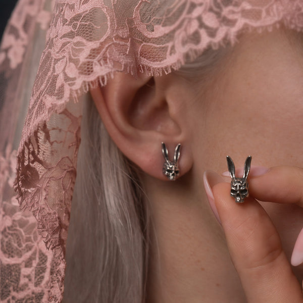 Darko Rabbit Skull stud earring worn on the ear while a second is held between fingers, pink lace veil behind