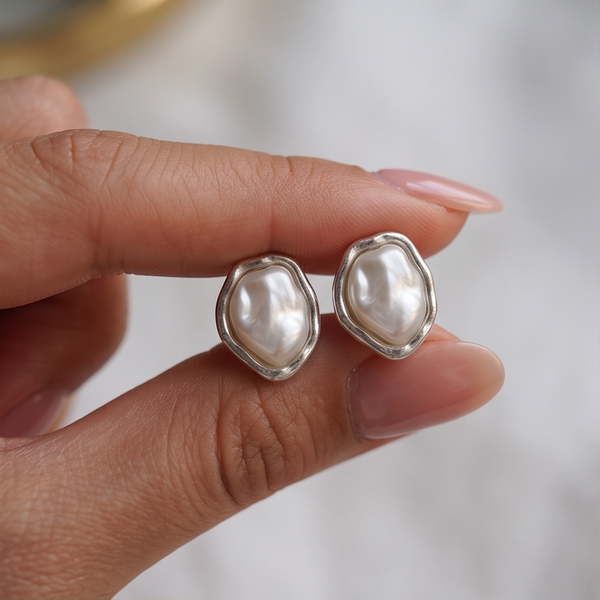 Baroque Pearl earrings held between fingertips, a pair with white pearls in wavy silver frames