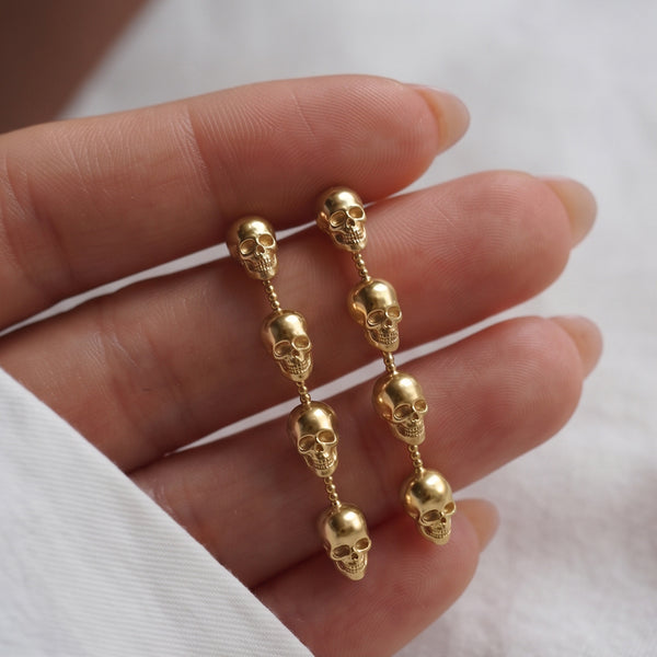 Skull Chain Drop earrings held in a hand, two gold drops of four skulls each on white fabric