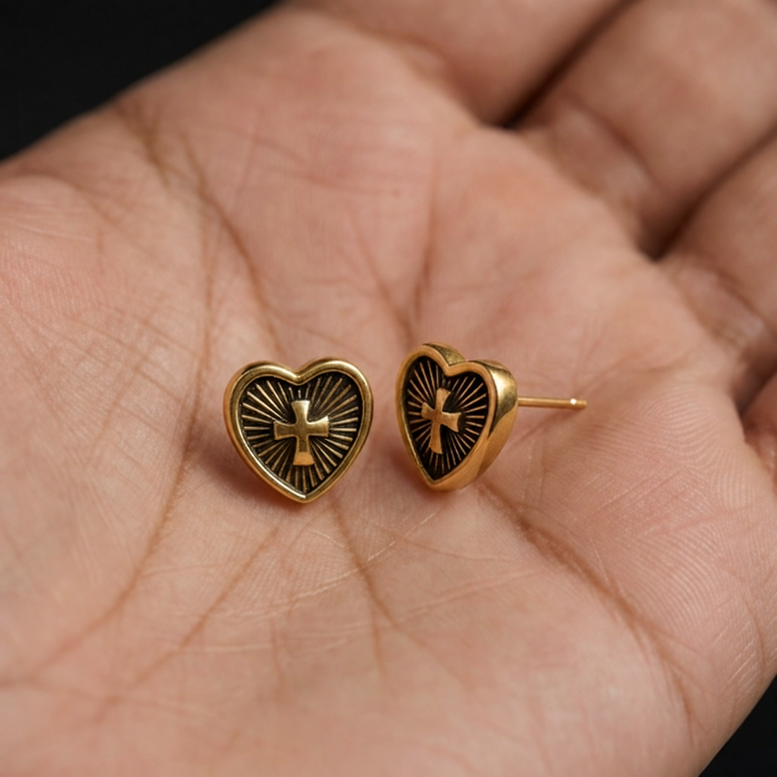 Heart cross stud earrings held on an open palm, gold hearts with a cross and ray details