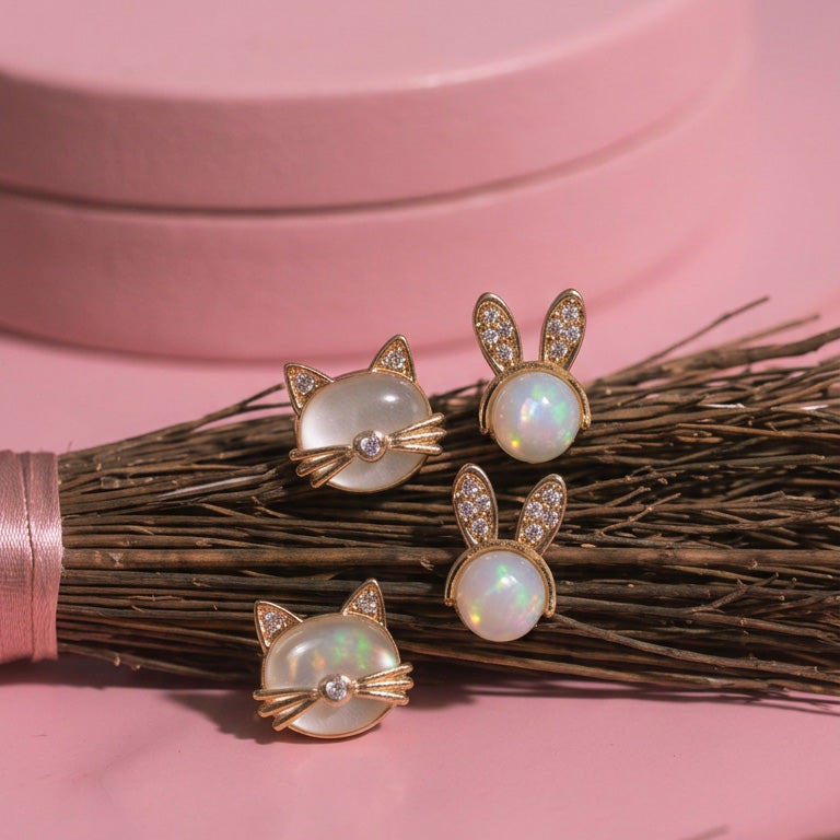 Mini Kitten stud earrings on a pink background beside a broom, with two bunny studs next to the cat faces