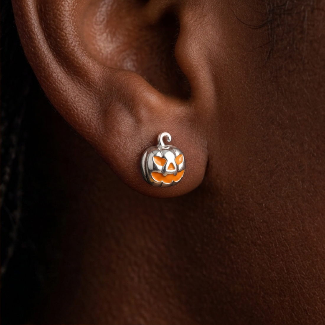 Silver Jack Pumpkin stud earrings worn on the ear, a silver jack-o'-lantern with an orange face