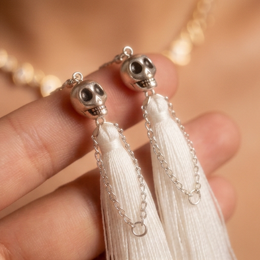 Skull Fringe Drop earrings, two silver skulls with white tassels held in a hand, close-up against bare skin