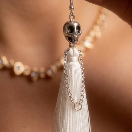 Skull Fringe Drop earrings, a silver skull with a long white tassel held up against bare skin and gold jewelry